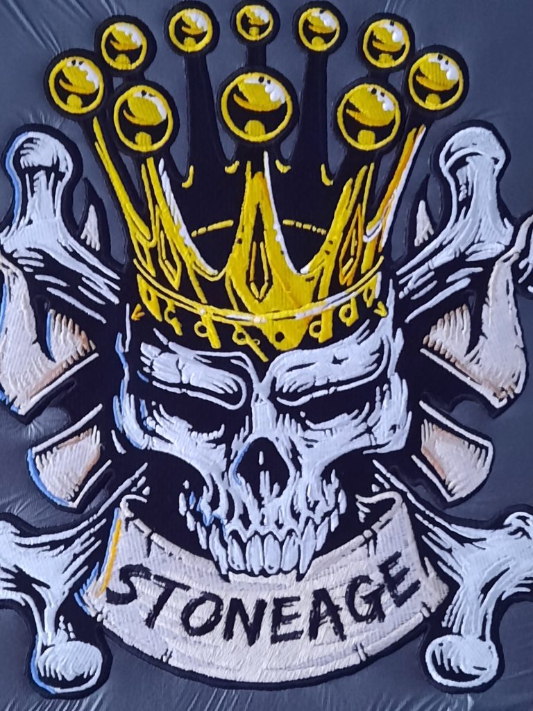Stickerei eines Totenschädels mit Krone. Im Mund Hält es ein Banner mit "Stoneage"