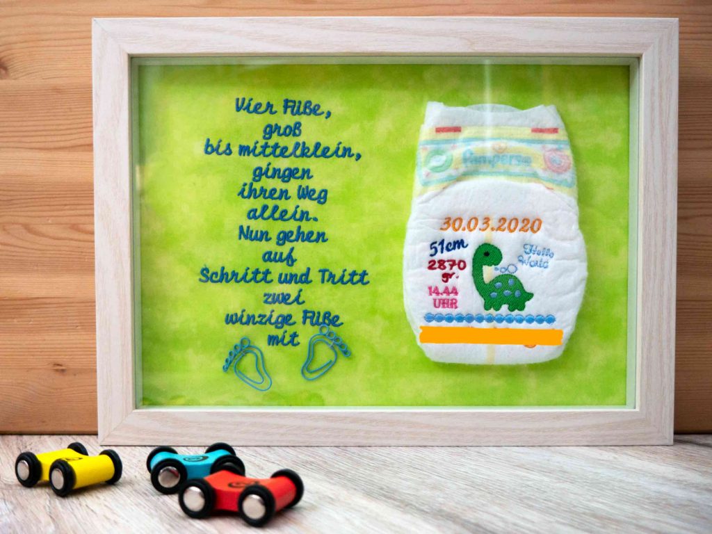 Windelbild im Holzrahmen. Der Hintergrund des Windelbilds ist ein grüner Stoff. Links ein gestickter Schriftzug: "Vier Füße groß bis mittelklein, gingen ihren Weg allein. Nun gehen auf Schritt und Tritt zwei winzige Füße mit". Die Windel ist rechts angeordnet. Darauf sind Geburtsdatum, -Größe, -Gewicht- und -Uhrzeit gestickt. Auch ein Kleiner grüner Dino, der "Hello World" sagt. Darunter ist der Name geschstickt- jedoch nicht zu sehen, da er von einem Balken verdeckt wird (Datenschutz)