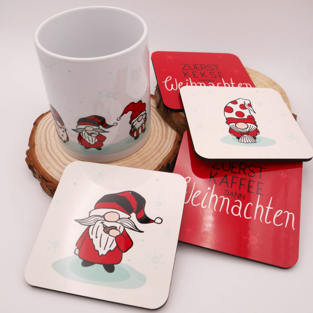 Das Bild zeigt eine weiße Tasse mit mehreren Weihnachts-Wichteln. Die Tasse steht auf einer runden Holzscheibe. Neben der Tasse liegen vier verschiedene quadratische Untersetzter, ebenfalls mit Weihnachtsmotiven. Ein Motiv zeigt einen Wichtel in rotem Mantel und schwarz-rot-geringelter Mütze, der einen Keks isst. Auf einem anderen Untersetzer ist ein Wichtel mit rot-gepunkteter Mütze. Der Hintergrund ist jeweils weiß mit Schneeflocken. Die beiden anderen Untersetzer haben einen roten Hintergrund. Auf dem einen steht "Zuerst Kekse, dann Weihnachten". Auf dem anderen der Spruch "Zuerst Kaffee, dann Weihnachten"