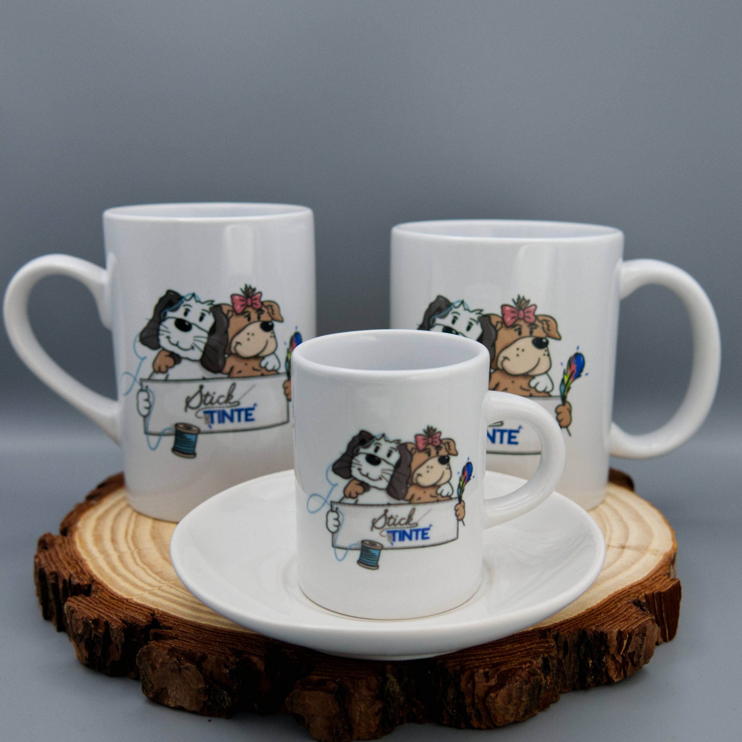 Das Bild zeigt drei Tassen in Unterschiedlicher Größe, die auf einer Holzscheibe stehen. Die vorderste Tasse ist eine Espressotasse. Die zwei Hinteren sind Becher in ebenfalls zwei verschiedenen Größen. Auf alles Tassen ist das Logo von "Stick und Tinte" aufgedruckt: Illustration von zwei Hunden, die ein Banner mit dem Logo von "Stick und Tinte" (stickundtinte.de) halten.