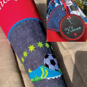 Schultüte in rot und antrazit. Im unteren Bereich Bestickt mit einem blau-grünem Fußballschuh. Im oberen Teil (im Foto abgeschnitten) mit dem Namen des Kindes. Ein Detailbild am oberen rechten Rand zeigt zwei Anhänger mit Einschulungsdatum und "1. Klasse"