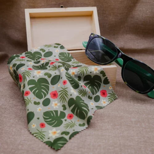 Micrfofastertücher für Brillen und Displays, mit Urban Jungle Muster: Monstera-Blätter, Blätter verschiedener Grünpflanzen und Dschungelblumen. Das Tuch liegt hier auf kleinen Schatzkiste. Es hängt halb herunter. An die Schatzkiste angelehnt ist auch eine grüne Sonnenbrille. Der Hintergrund ist brauner Leinenstoff