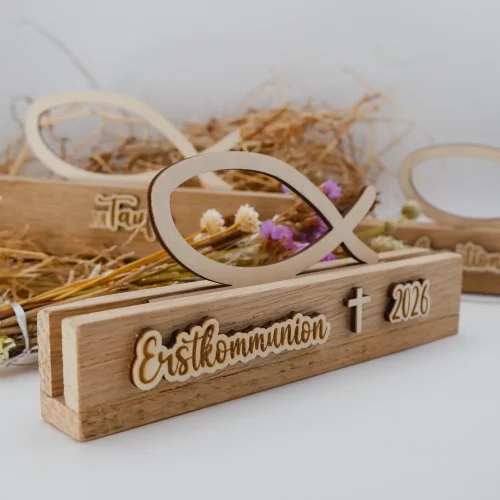 Schöne Tischdeko zur Erstkommunion Fisch aus Holz, bestehend aus einem Aufsteller und dem Fisch-Einschieber. Der Aufsteller ist beschriftet mit Erstkommunion, Kreuz und Jahreszahl (hier 2026). Die Beschriftung ist ebenfalls aus Holz. Dadurch gibt es einen schönen 3D-Effekt. Das Foto zeigt die Ansicht schräg von der Seite. Im Hintergrund sind verschwommen je eine Variante für Taufe und für Kommunion zu erkennen.