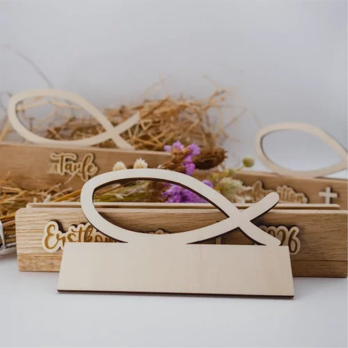 Schöne Tischdeko für christliche Feste. Der Einschieber Fisch kann in die Klötzchen-Aufsteller eingesetzt werden. Der Fisch aus Holz lehnt an einem Klötzchenaufsteller für Erstkommunion. Im Hintergrund sind verschwommen je eine Variante für Taufe und für Kommunion zu erkennen.