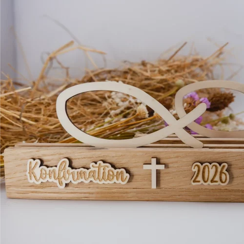 Schöne Tischdeko zur Konfirmation mit Fisch aus Holz, bestehend aus einem Aufsteller und dem Fisch-Einschieber. Der Aufsteller ist beschriftet mit Konfirmation, Kreuz und Jahreszahl (hier 2026). Die Beschriftung ist ebenfalls aus Holz. Dadurch gibt es einen schönen 3D-Effekt. Das Foto zeigt die Ansicht von Vorne in näherer Aufnahme Im Hintergrund ist ein weiterer Fisch zu erkennen, sowie Stroh und Trockenblumen