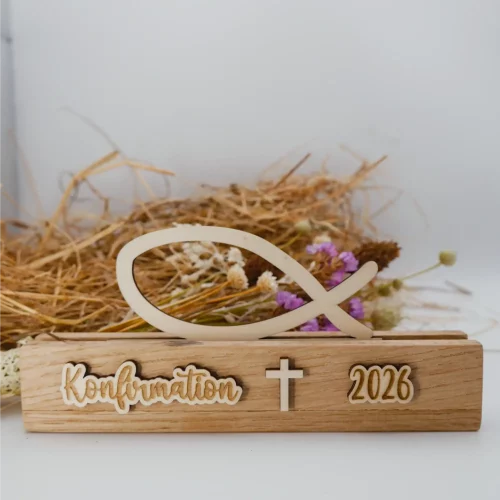 Schöne Tischdeko zur Konfirmation mit Fisch aus Holz, bestehend aus einem Aufsteller und dem Fisch-Einschieber. Der Aufsteller ist beschriftet mit Konfirmation, Kreuz und Jahreszahl (hier 2026). Die Beschriftung ist ebenfalls aus Holz. Dadurch gibt es einen schönen 3D-Effekt. Das Foto zeigt die Ansicht von Vorne. Im Hintergrund ist ein weiterer Fisch zu erkennen, sowie Stroh und Trockenblumen
