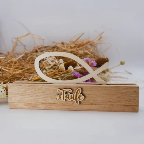 Schöne Tischdeko zur Taufe mit Fisch aus Holz, bestehend aus einem Aufsteller und dem Fisch-Einschieber. Der Aufsteller ist beschriftet mit "zur Taufe". Die Beschriftung ist ebenfalls aus Holz. Dadurch gibt es einen schönen 3D-Effekt. Das Foto zeigt die Ansicht von Vorne. Die Taufdeko steht vor Stroh und Trockenblumen.
